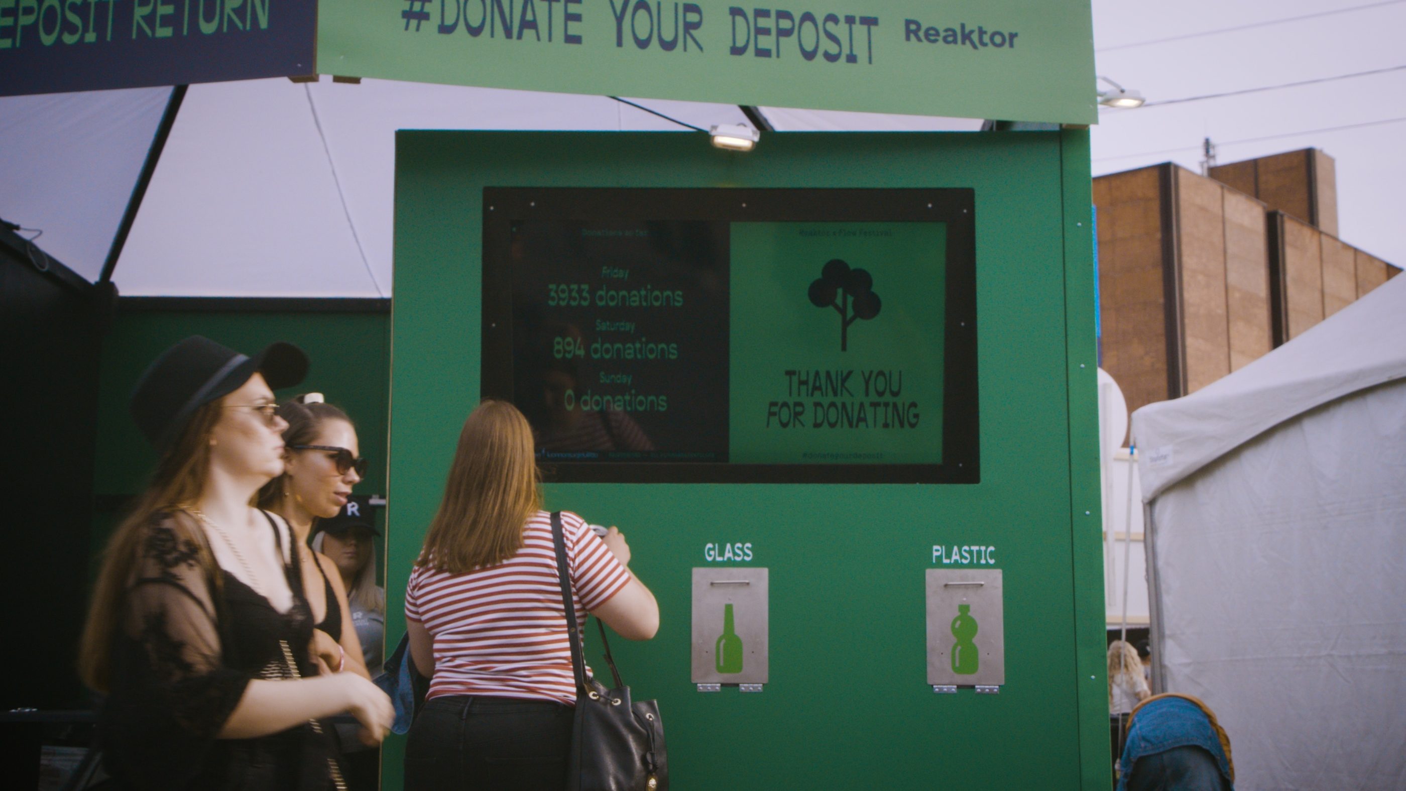 A deposit kiosk on the festival area: Two women are walking past the kiosk in summer clothes. One person is depositing a bottle to the kiosk. A screen on the kiosk reads: Thank you for donating, with an image of a tree.