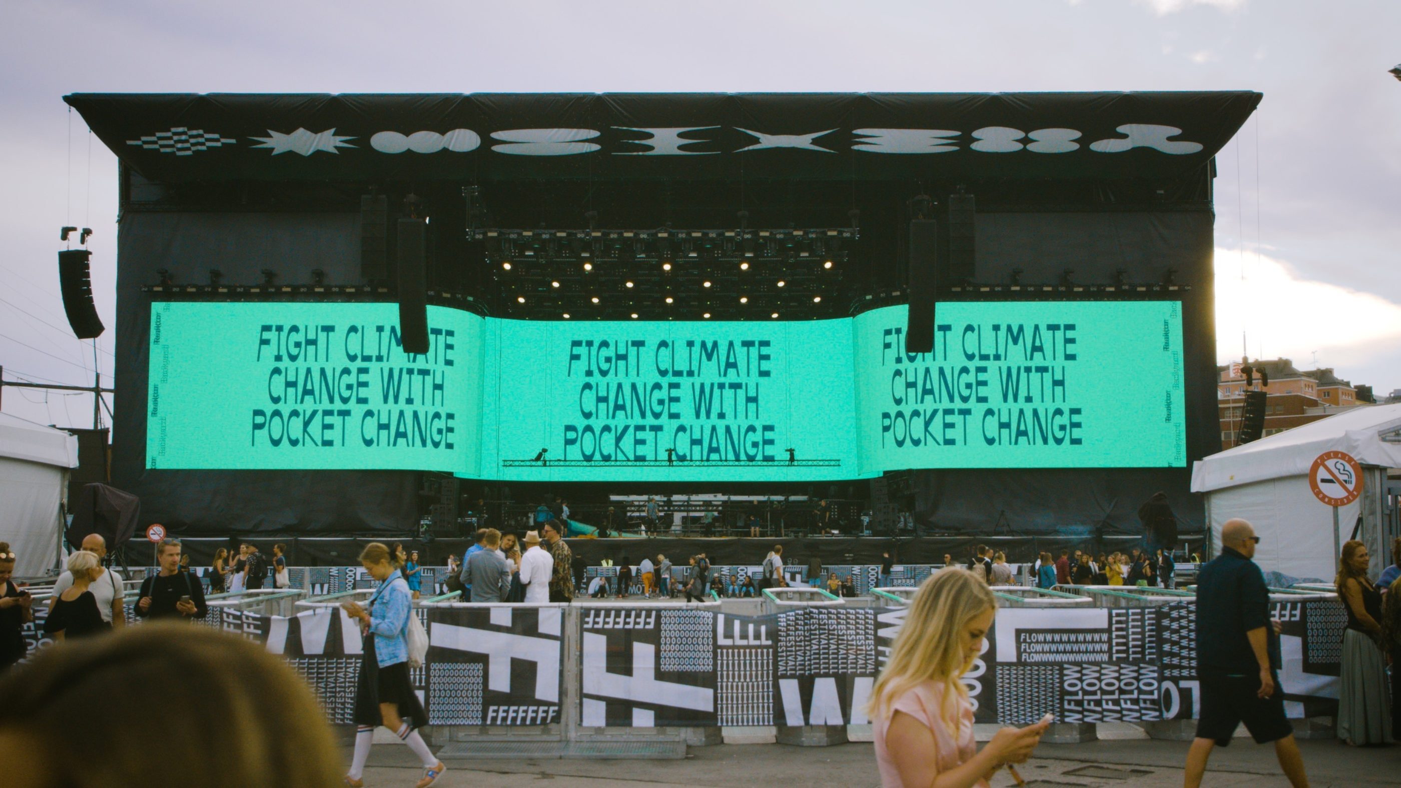 An image of a festival stage with a small crowd gathered in front of it and individual people walking past it. The screens on the stage read: Fight climate change with pocket change.