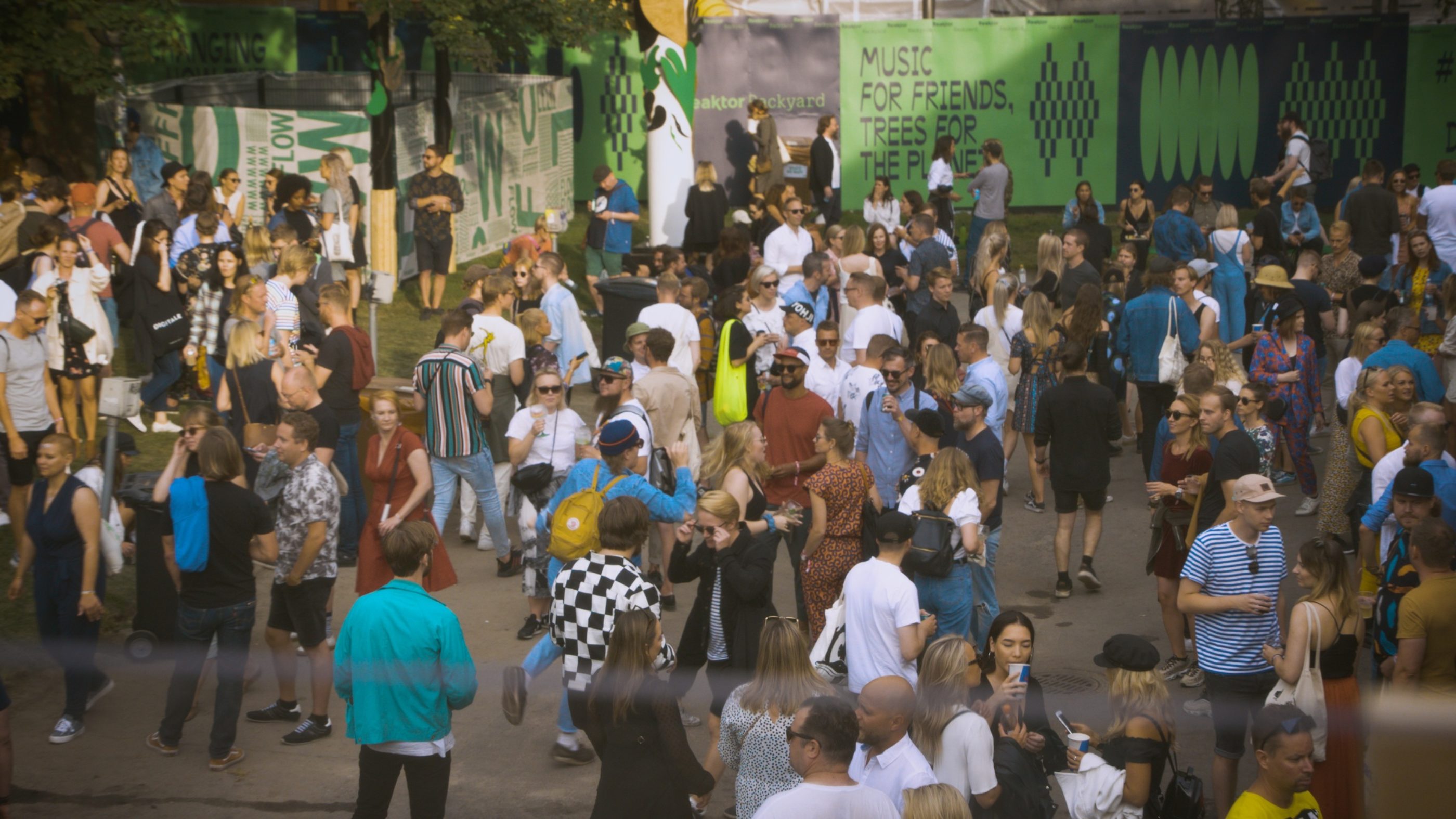 A festival crowd in summer clothes partying and talking amongst themselves. A wall on the background reads: Music for friends, trees for the planet. 