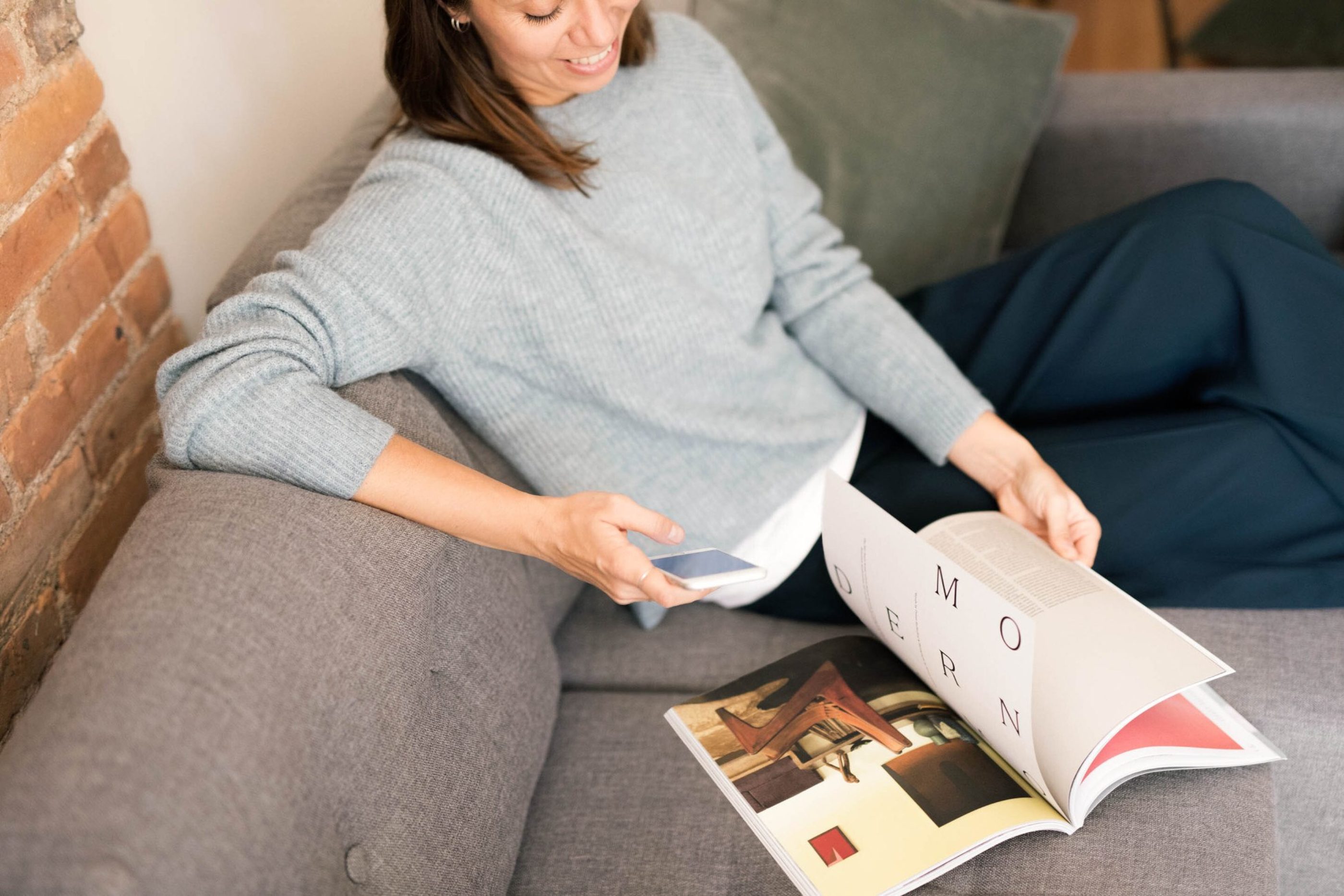 A brown-haired woman sitting on couch, browsing a magazine while holding a smart phone on her hand.